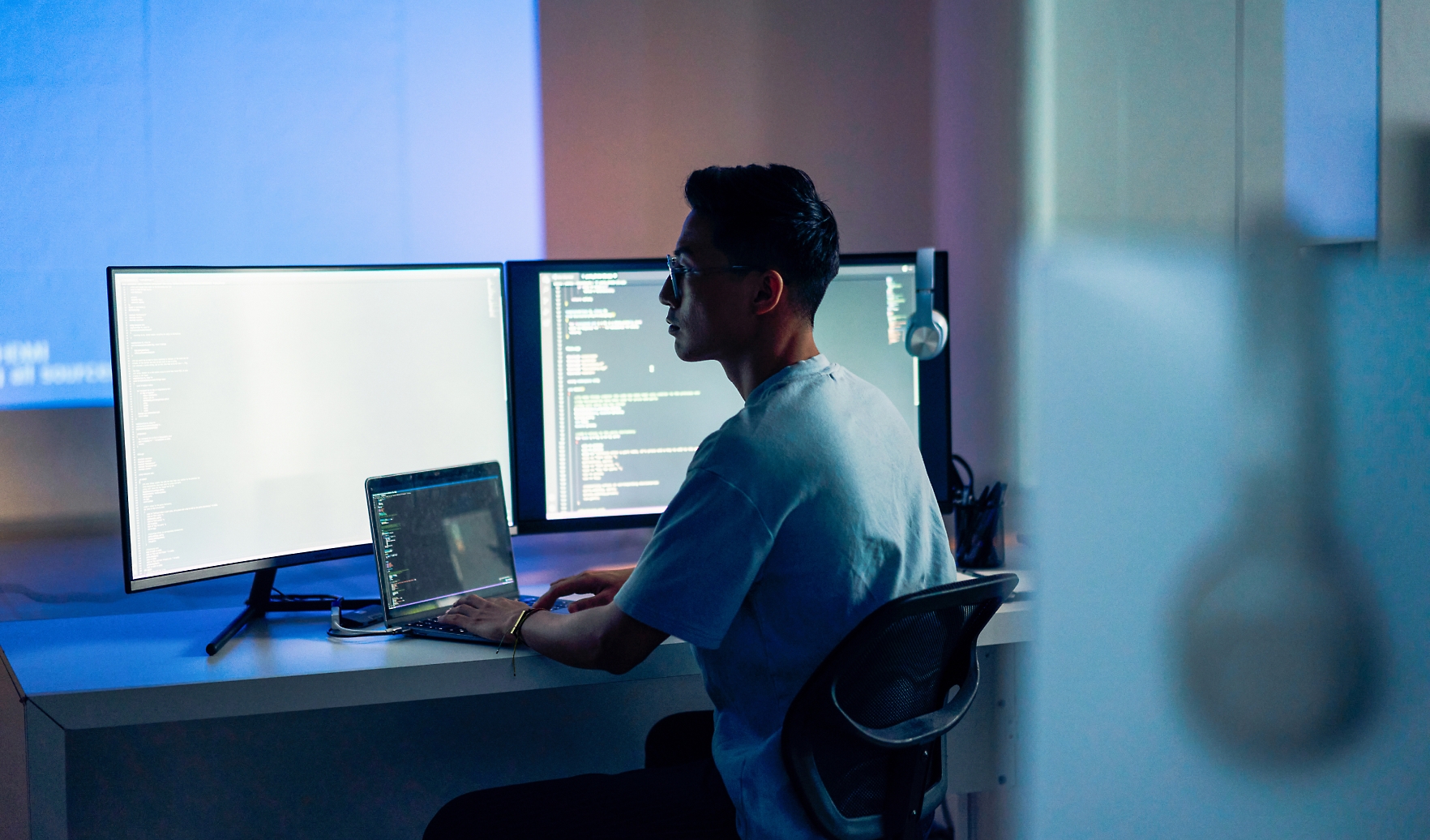 Person sitting at a desk working on a laptop, with two monitors displaying code in a dimly lit room