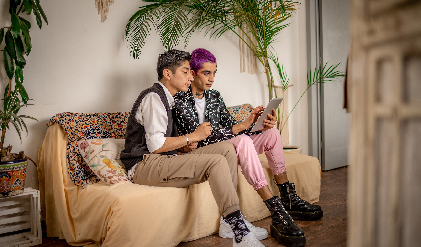 Two people sitting together on a couch, looking at a tablet. One has short gray hair and wears beige pants