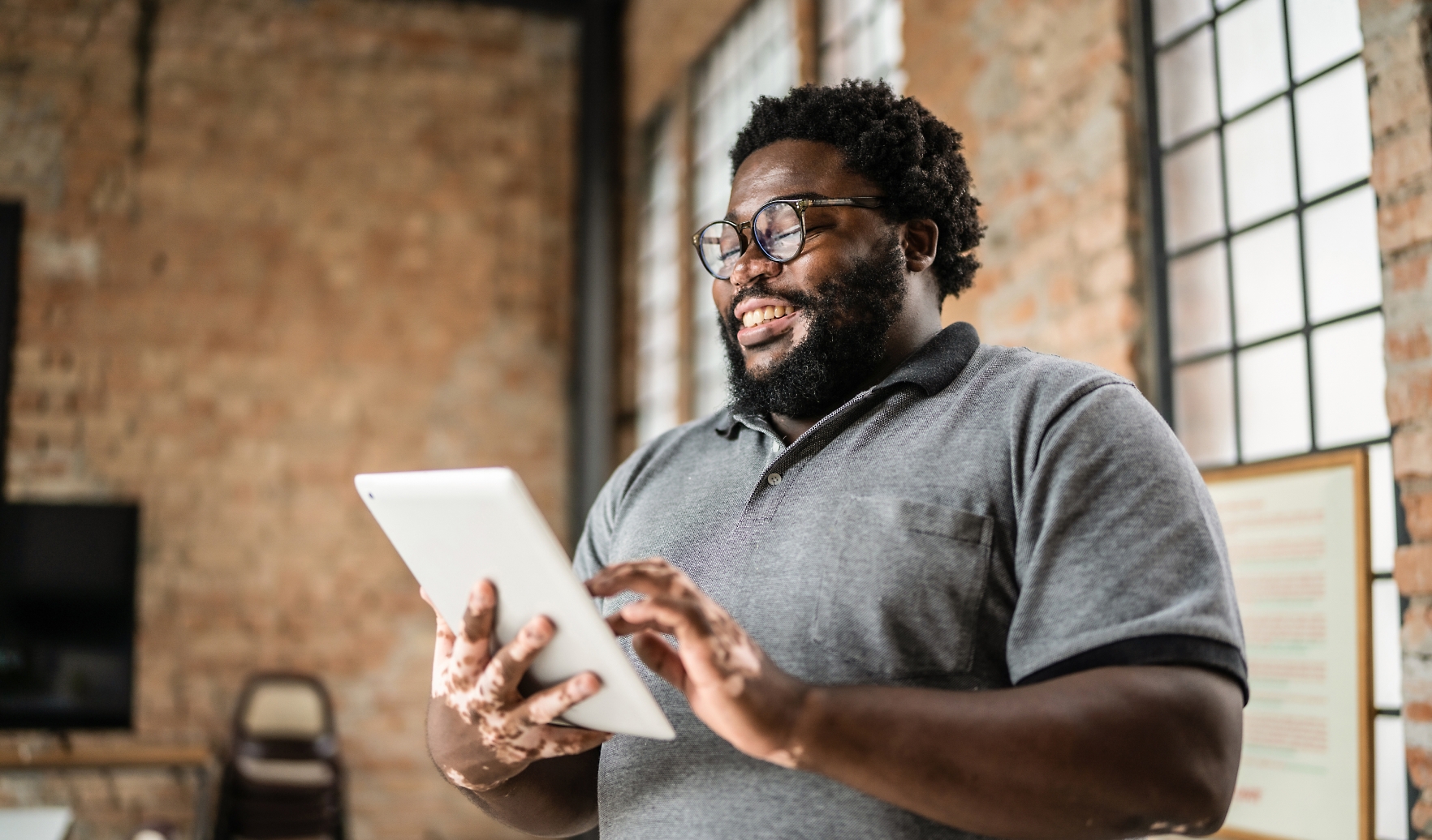 A person wearing glasses and a grey polo shirt is smiling while using a tablet in a room