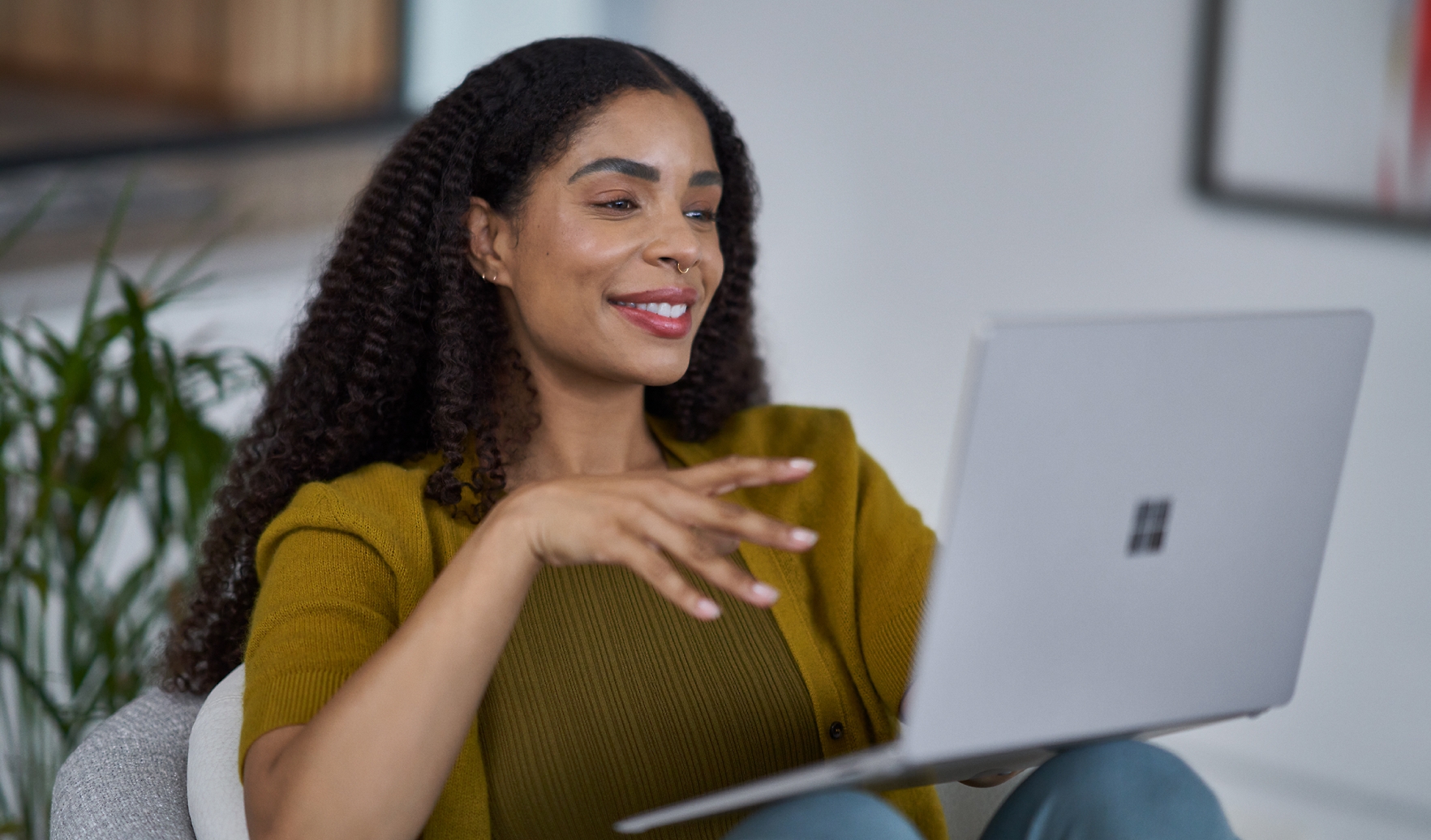 A woman with curly hair and wearing a yellow cardigan smiles while using a laptop