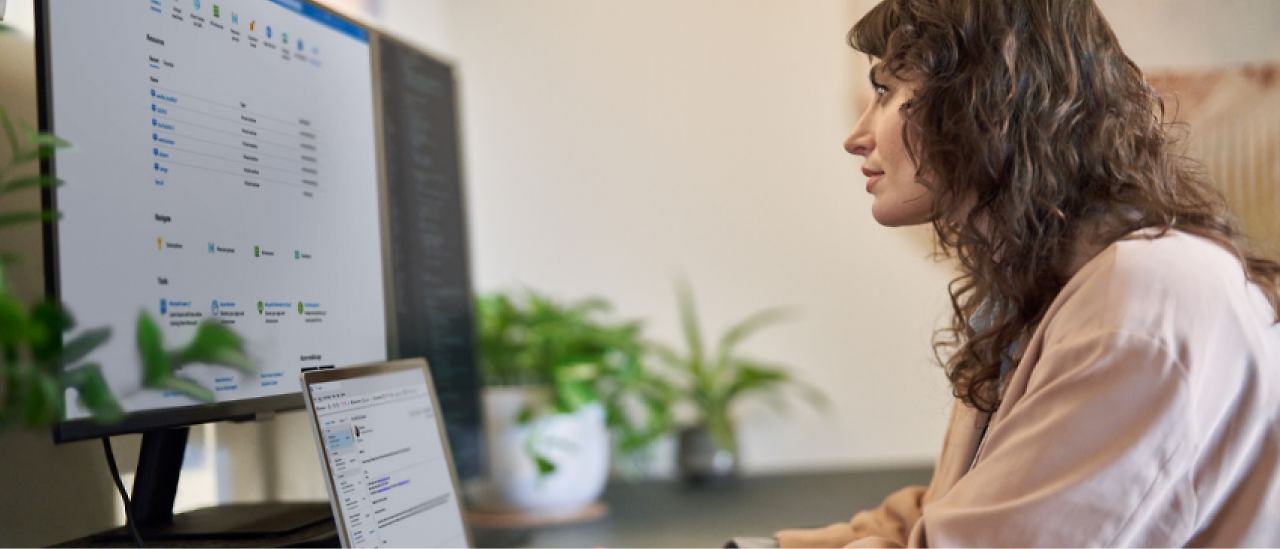 A woman with curly hair sits at a desk working on a desktop computer and a laptop. 