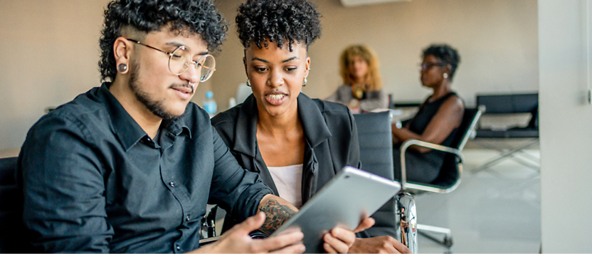 Two professionals discussing over a digital tablet in a modern office setting, with another person 