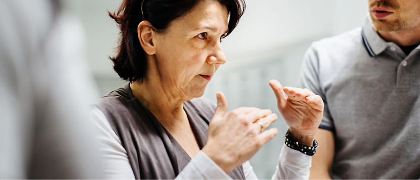 A middle-aged woman gestures while speaking intently to a man, partially visible, in an office setting.