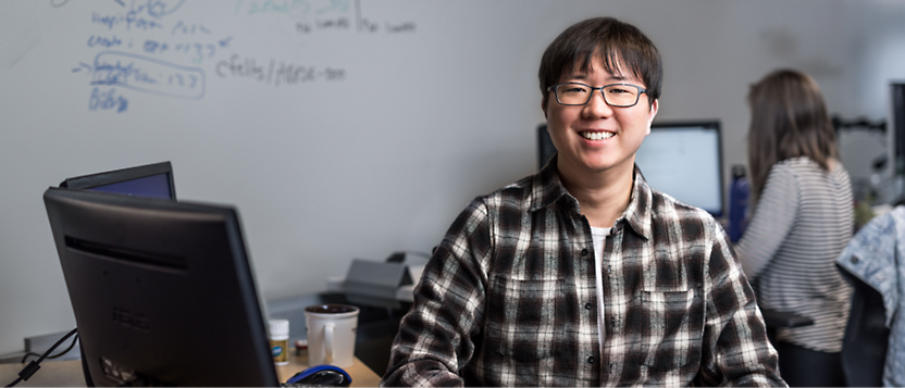 A smiling East Asian person wearing glasses and a plaid shirt seated in front of a computer in an office with a whiteboard 