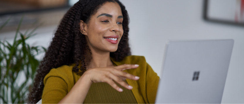 A woman with curly hair smiles gently while looking at her laptop screen, wearing a mustard yellow sweater.