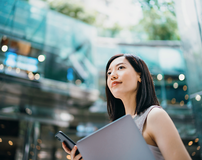 Eine Frau hält ein Smartphone und einen Laptop und steht im Freien vor einem modernen Glasgebäude im Hintergrund.