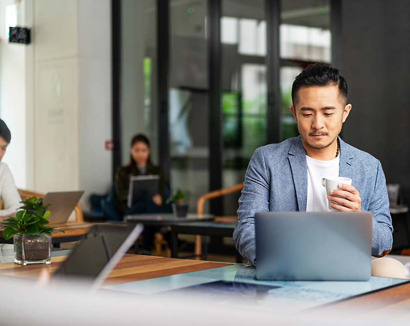 A man in a blue blazer sits at a desk using a laptop while holding a coffee cup