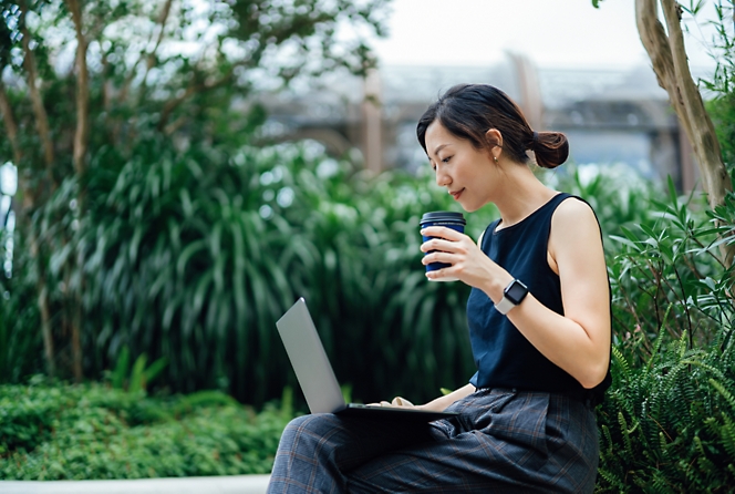 Una mujer se encuentra al aire libre en un banco con hojas detrás de ella, sosteniendo una taza de café en una mano y usando un portátil