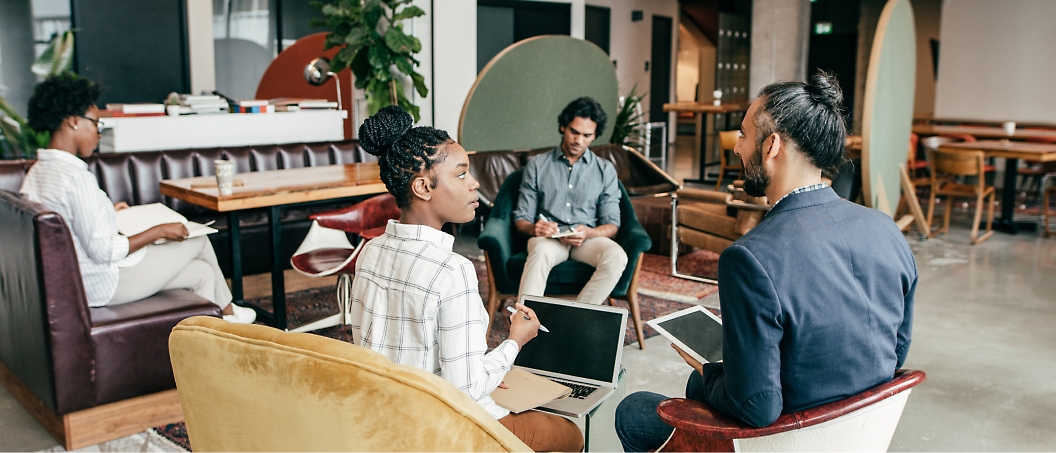 Four individuals are seated in a modern office lounge, engaged in conversation and working on laptops and notebooks.