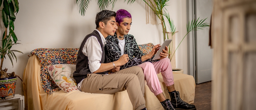 Two people sitting together on a couch, looking at a tablet. One has short gray hair and wears beige pants