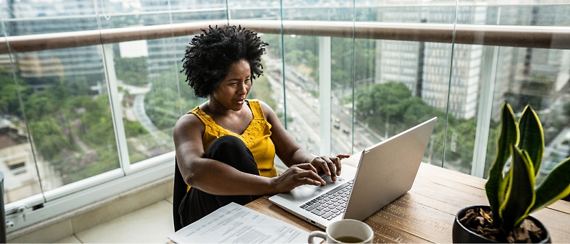 Eine Frau sitzt auf einem Balkon mit Blick auf die Stadt und arbeitet an einem Laptop. Eine Pflanze, ein Kaffeebecher und Papiere