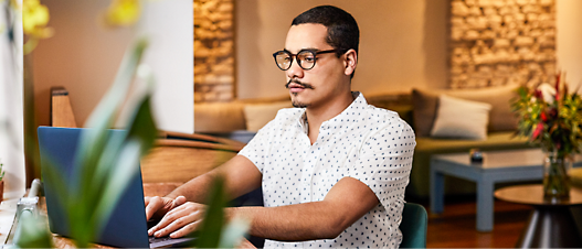 A man wearing glasses working intently on a laptop in a cozy, well-decorated room with stone walls and a floral arrangement.