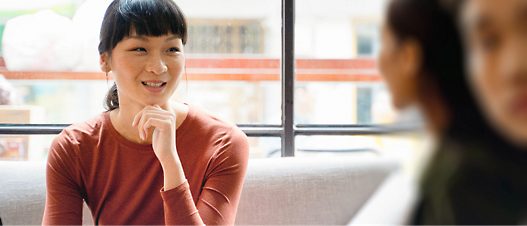 A smiling asian woman in a red top resting her chin on her hand, sitting in a brightly lit café