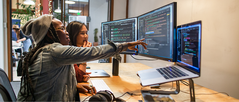 Two people sitting at a desk working on multiple monitors displaying code, one person pointing at a screen.