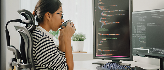 A woman in a wheelchair sips coffee while looking at computer screens displaying code in a bright office.