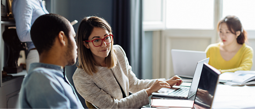 Two professionals discussing over a laptop in an office setting while a colleague works in the background.