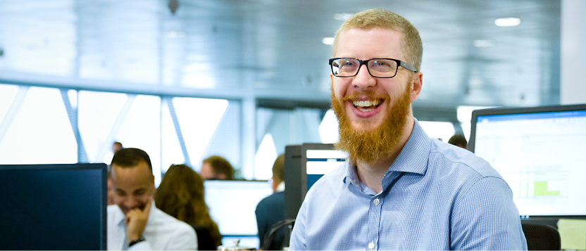 A man with a red beard and glasses smiles while sitting at his desk in a busy office environment with colleagues 