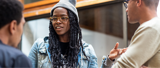 Three young adults engaged in conversation outdoors, with a woman in a denim jacket and glasses 