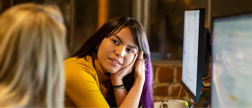 Two people are sitting in an office, with one person looking attentively at the other. They are seated at desks