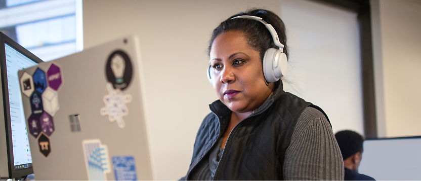 A woman wearing headphones works on a computer in an office. The laptop has several stickers on the back.