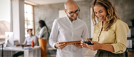 Two professionals, a bald man and a blonde woman, smile while observing a tablet, in a modern office with coworkers.