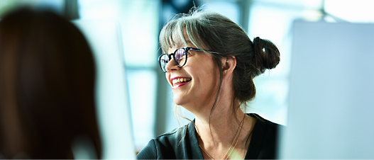 Smiling middle-aged woman with glasses and a bun discussing with a colleague in a bright office setting.