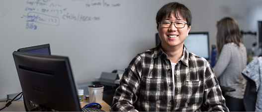 A smiling person with glasses wearing a plaid shirt, sitting at a desk with a computer monitor, in an office with a whiteboard