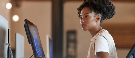 A young woman working attentively at a standing desk with computer monitors in a modern office setting.