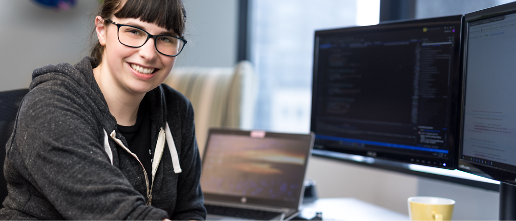 A person wearing glasses and a hoodie sits at a desk with a laptop and two monitors displaying code