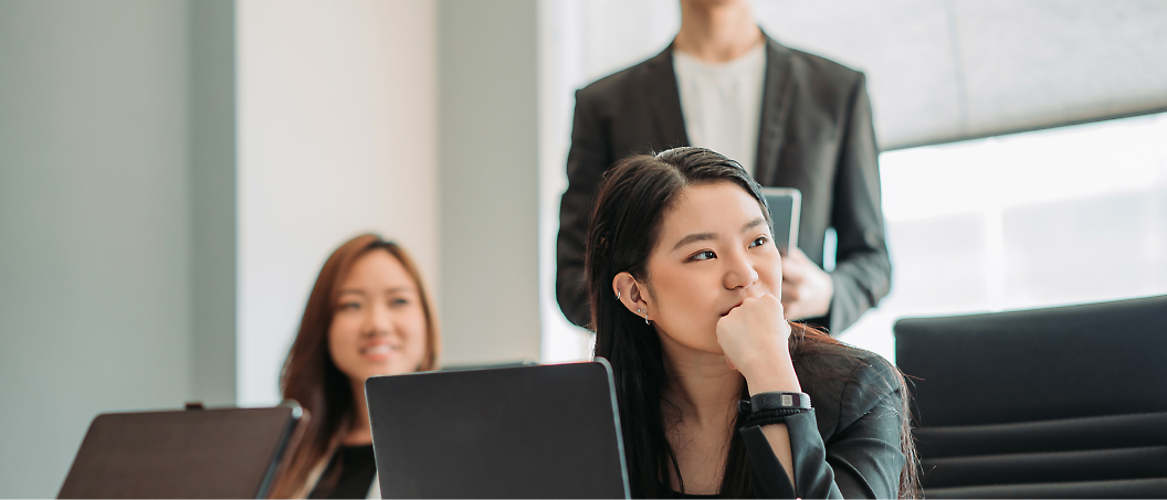 Three people in a meeting room with laptops; one woman sits and looks thoughtfully, a second woman smiles