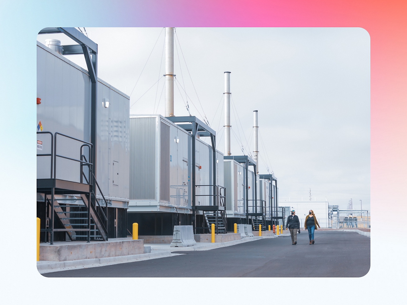 Due persone che camminano vicino a una fila di container industriali con alti camini in uno stabilimento sotto un cielo nuvoloso.