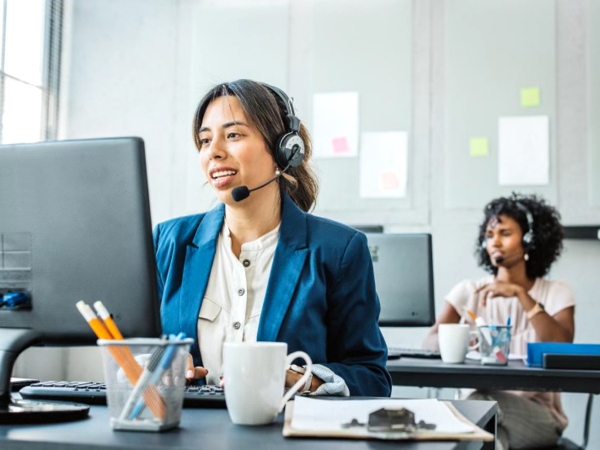 A customer service representative wearing a headset and smiling while working at her computer in an office setting.