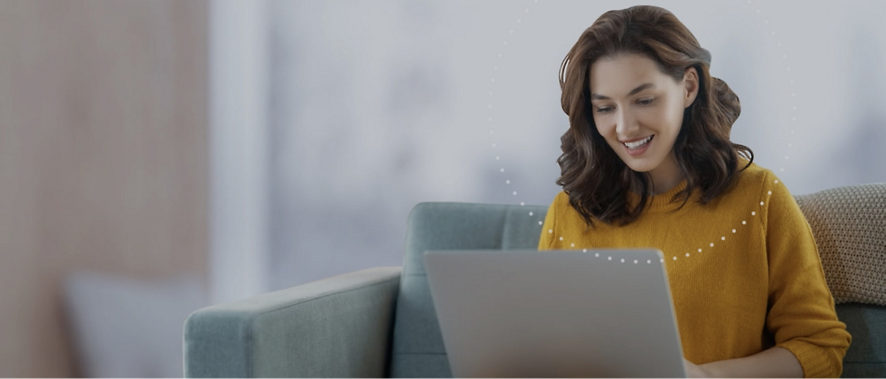 A person, sitting on a sofa, smiles while using a laptop. The background is blurred.