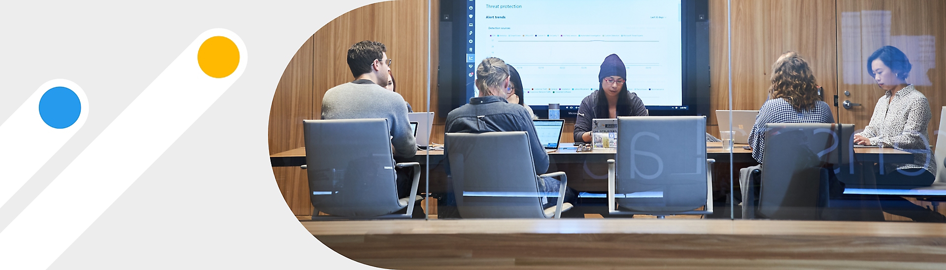 A meeting with 7 people in a conference room with a large screen on the back wall displaying notes in Teams.