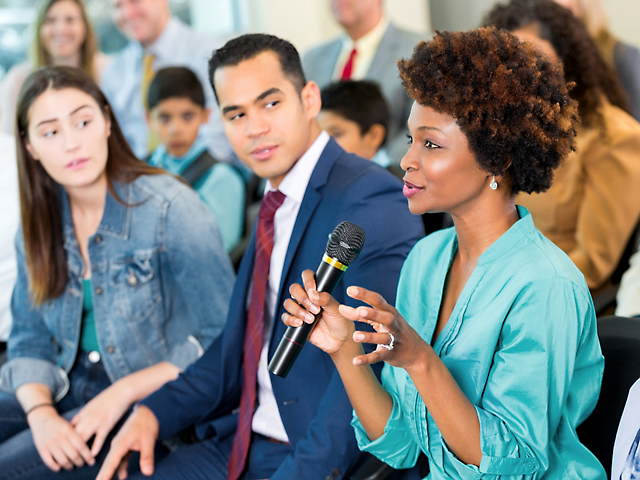 Een vrouw spreekt in een microfoon tijdens een discussie op een conferentie, met aandachtige toehoorders op de achtergrond.