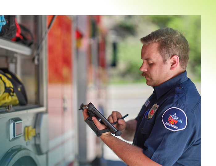 Пожарный в синей форме со значком пишет на планшете рядом с пожарной машиной.