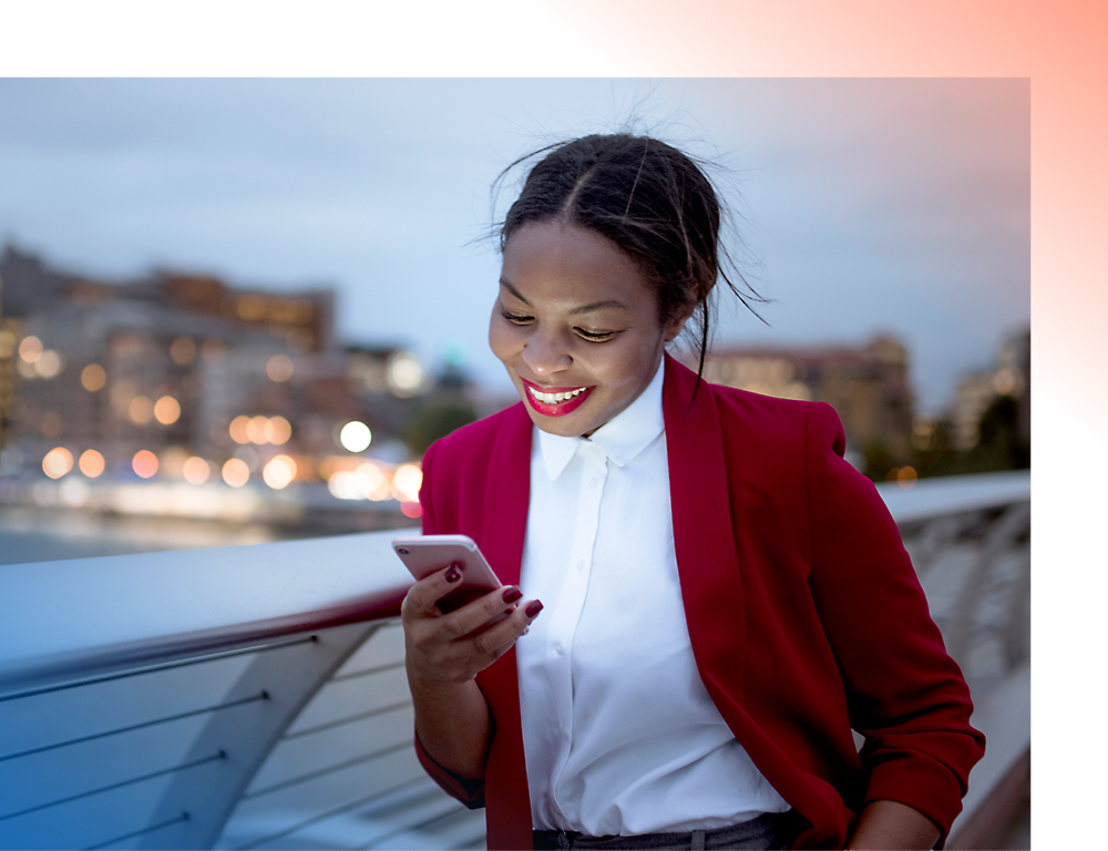 Una mujer sonriente con una chaqueta roja usando un smartphone en un puente de ciudad al atardecer.