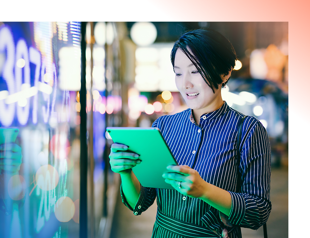 A woman with short hair using a tablet, stands by a brightly lit digital display in an urban setting at night.