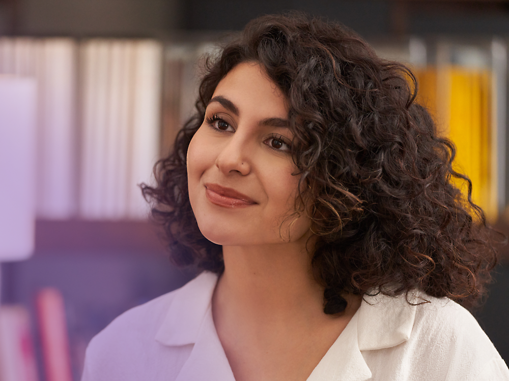 A woman with curly hair smiling gently, wearing a light pink blazer, with a softly blurred background.