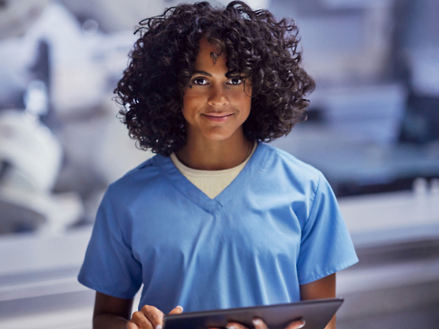 A person wearing a blue scrub, holding a tablet and smiling.