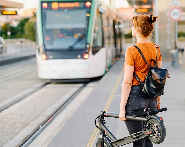 A person with a folded electric scooter and a backpack waits on the sidewalk as a tram approaches.