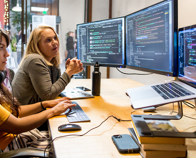 Two women working and discussing with multiple monitors on the desk