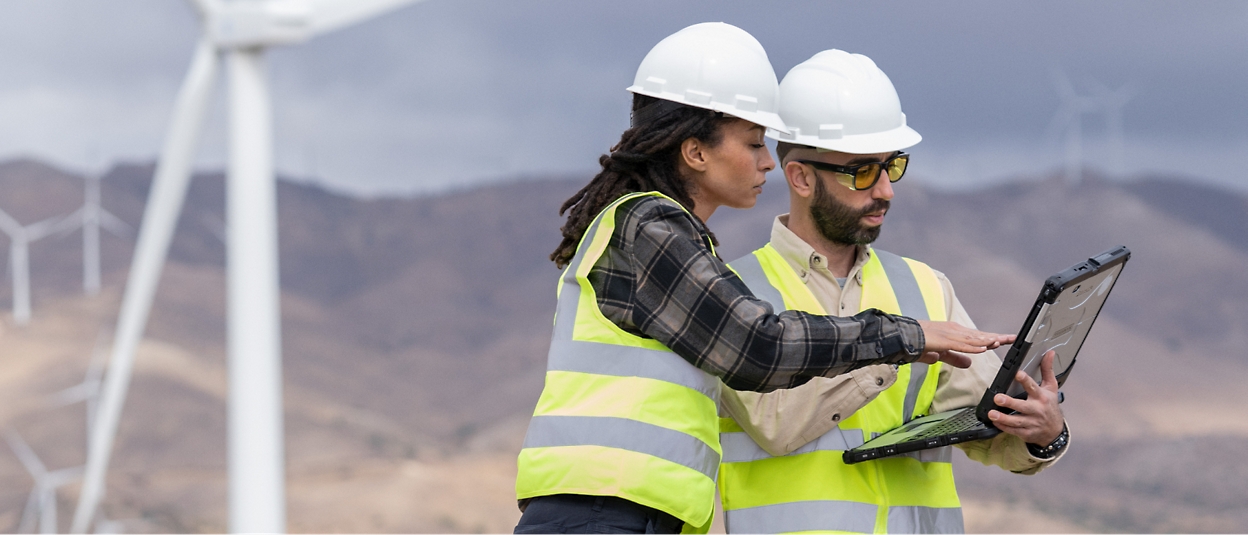 Dos trabajadores con casco y chaleco reflectante examinan un ordenador portátil cerca de unas turbinas eólicas en un paisaje montañoso.