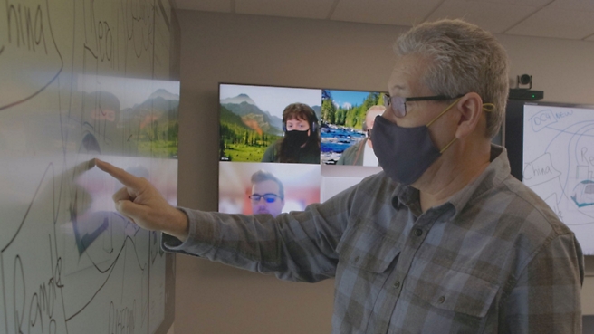 A man wearing a face mask is pointing at a screen.