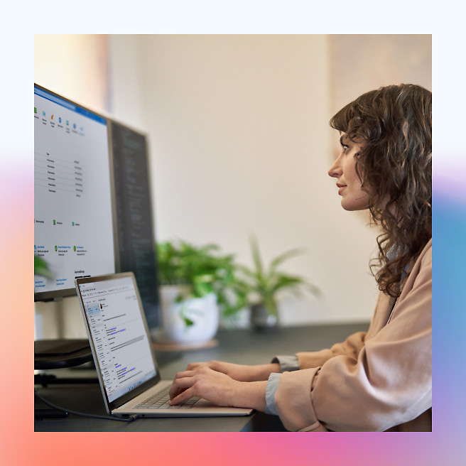 A woman working on a laptop with a larger monitor next to it in an office setting.