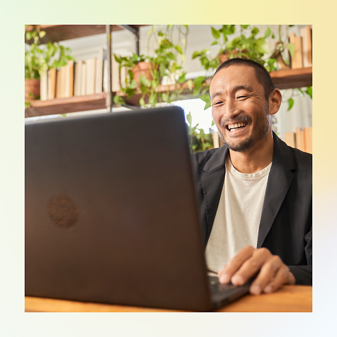 Man smiling while using a laptop in a room with plants and books.