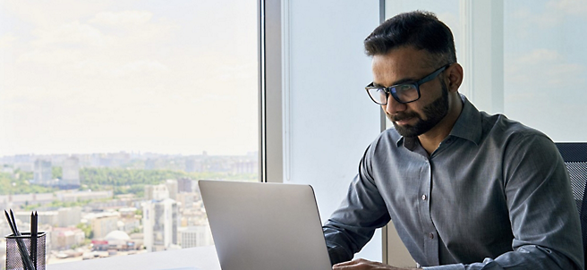 A man with glasses working on a laptop in an office with city views visible through large windows.