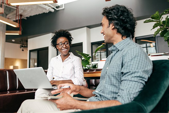 One person and another sit at a table, conversing while one uses a laptop