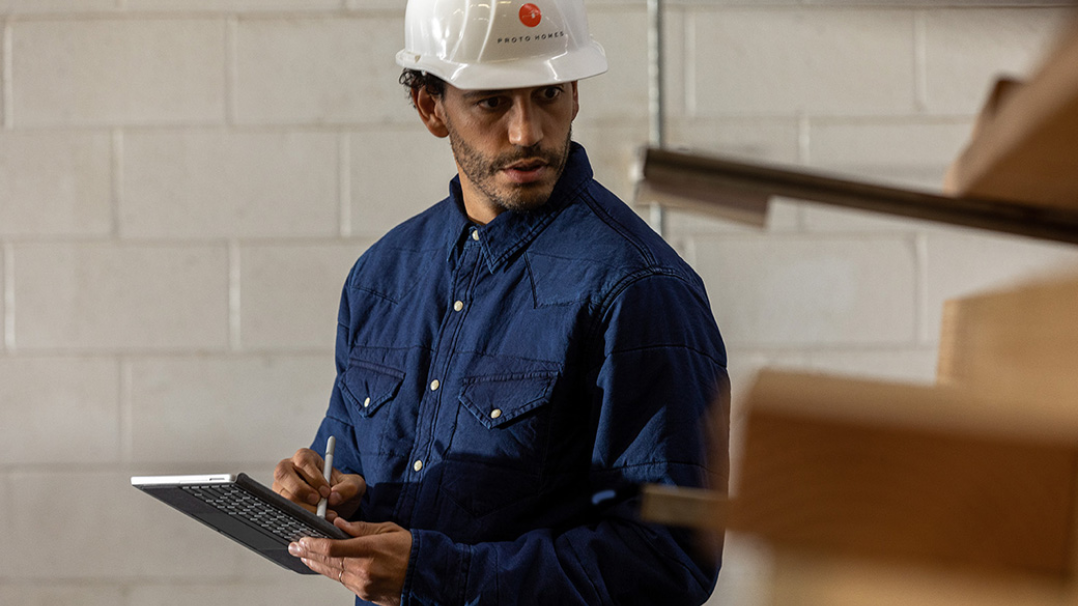 Se muestra un trabajador con un casco en una zona industrial que sostiene un dispositivo Surface Go 2 en modo tableta