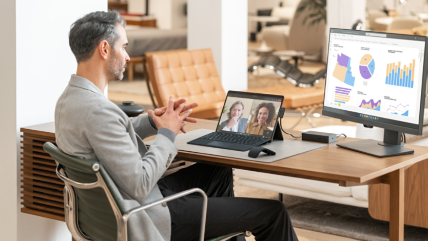 A man sitting at a desk video conferencing with two people on a 2-in-1 PC, and a computer monitor showing digital graphs and pie charts